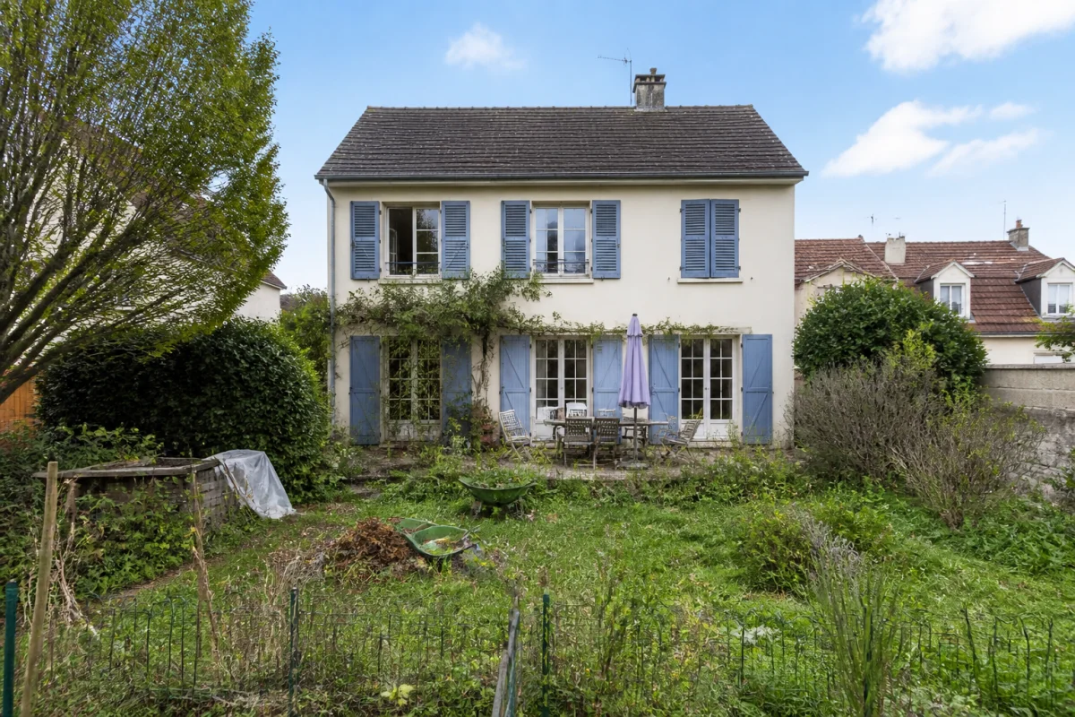 Maison à vendre avec jardin négligé, pelouse haute, terrasse encombrée et façade nécessitant une préparation avant vente